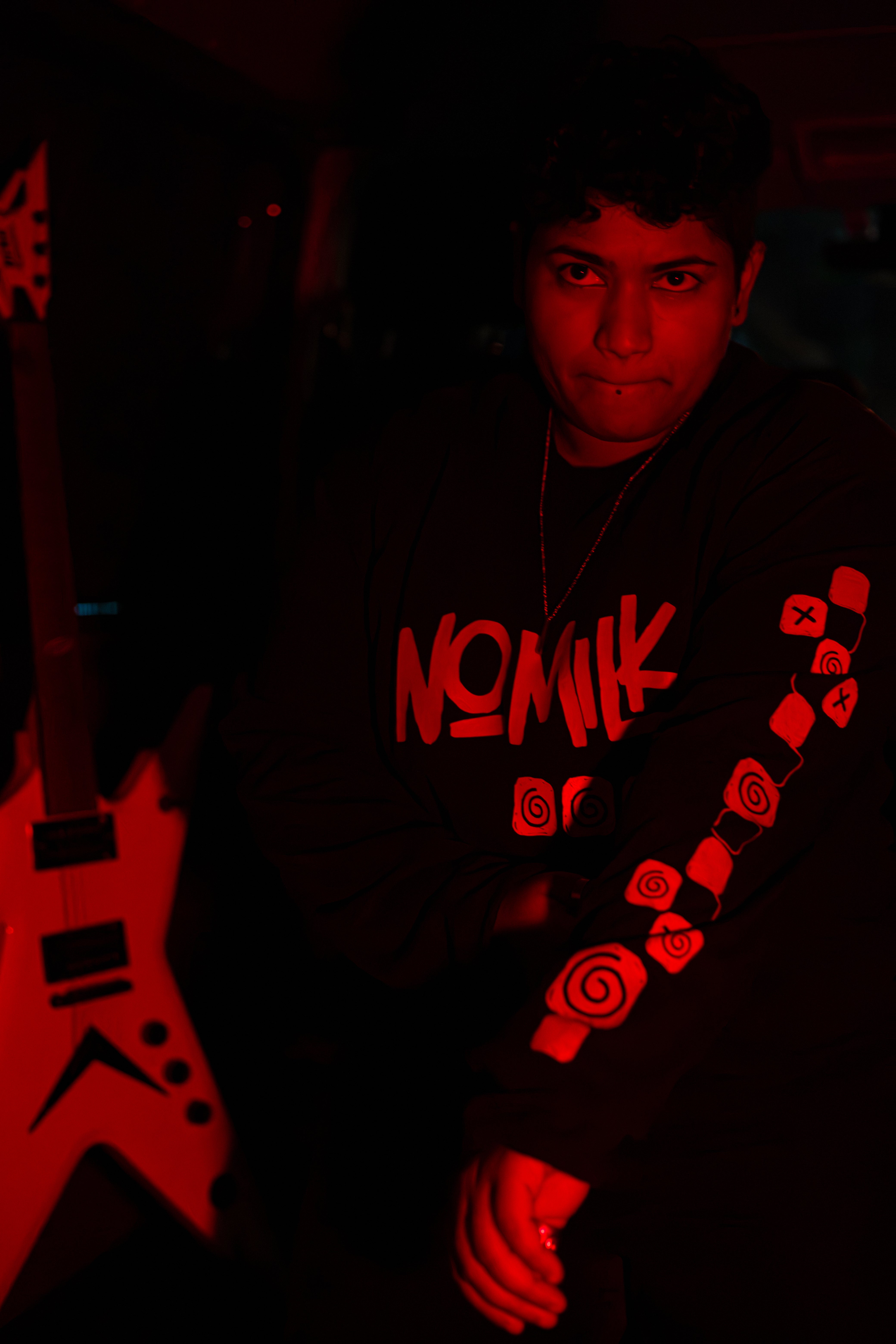 Featuring Noz, in the Noughts Win shirt, showing the sleeve under a red light, electric guitar behind her.